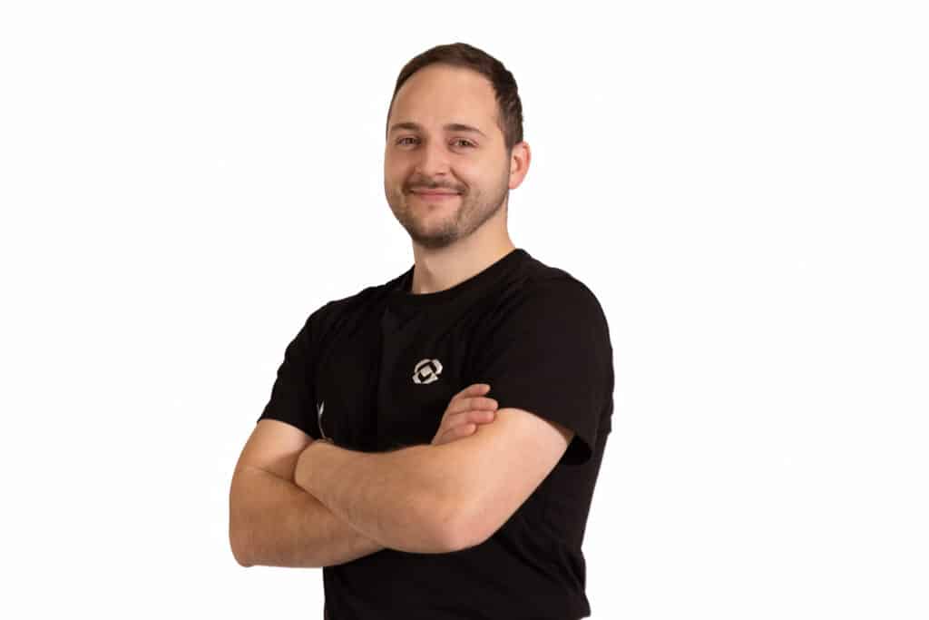 Dominik Kramár z KRAMÁR kuchynského štúdia – portrét s priateľským úsmevom, v čiernom firemnom tričku, na bielom pozadí.