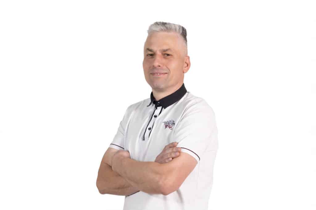 Portrét Františka Kováčika, muža s krátkymi šedivými vlasmi v bielom tričku s čiernym golierom, s prekríženými rukami, na bielom pozadí.