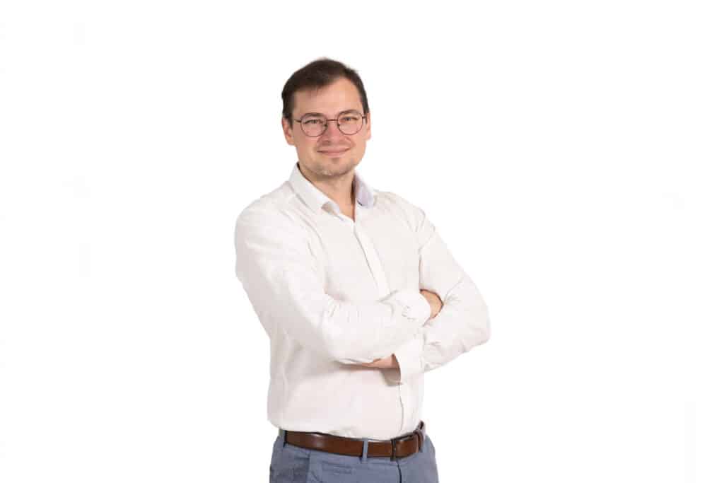 Portrét Lukáša Peška, muža s okuliarmi v bielej košeli s prekríženými rukami, na bielom pozadí.